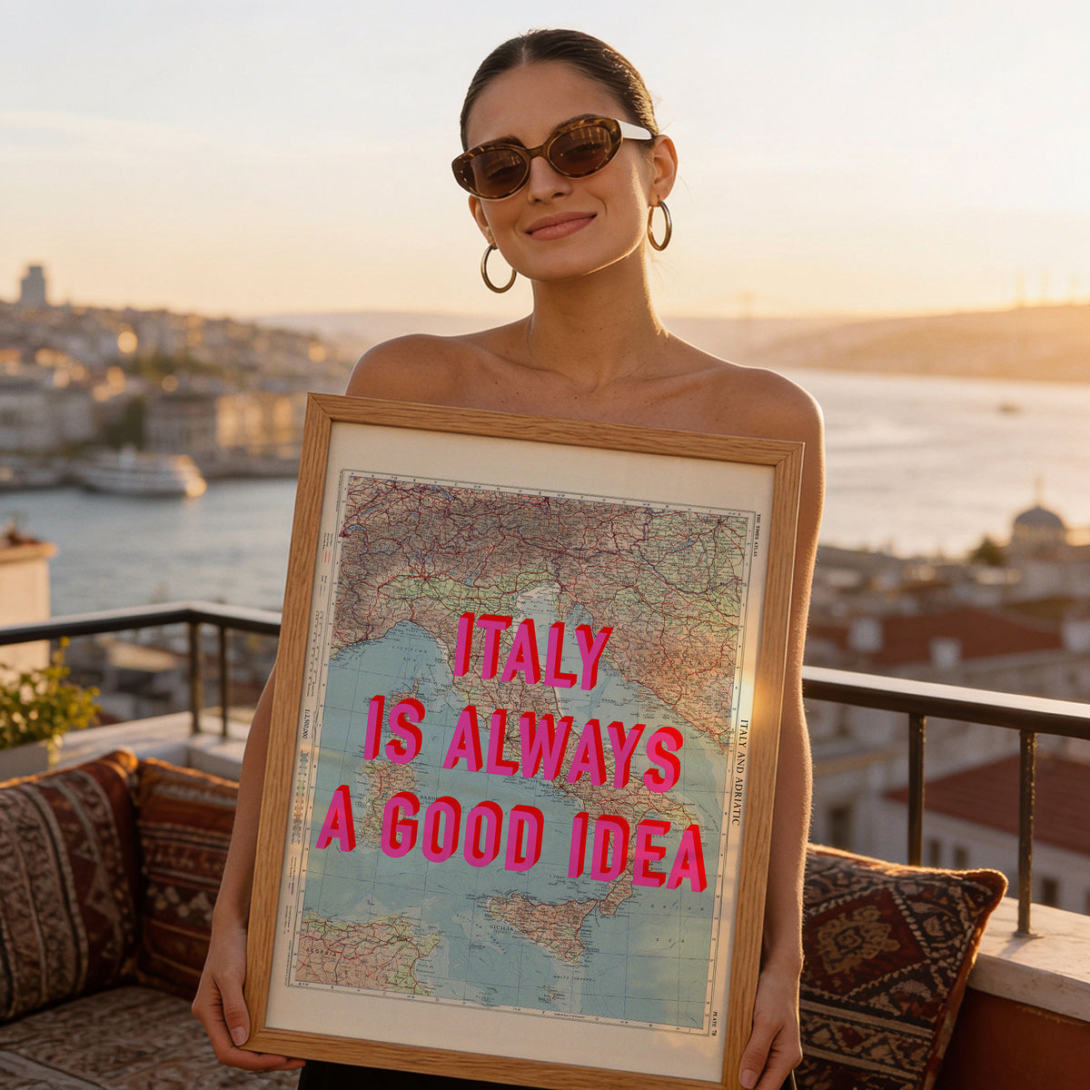 Woman holding a framed Italy Is Always A Good Idea pop art map print on a balcony overlooking the sea at sunset